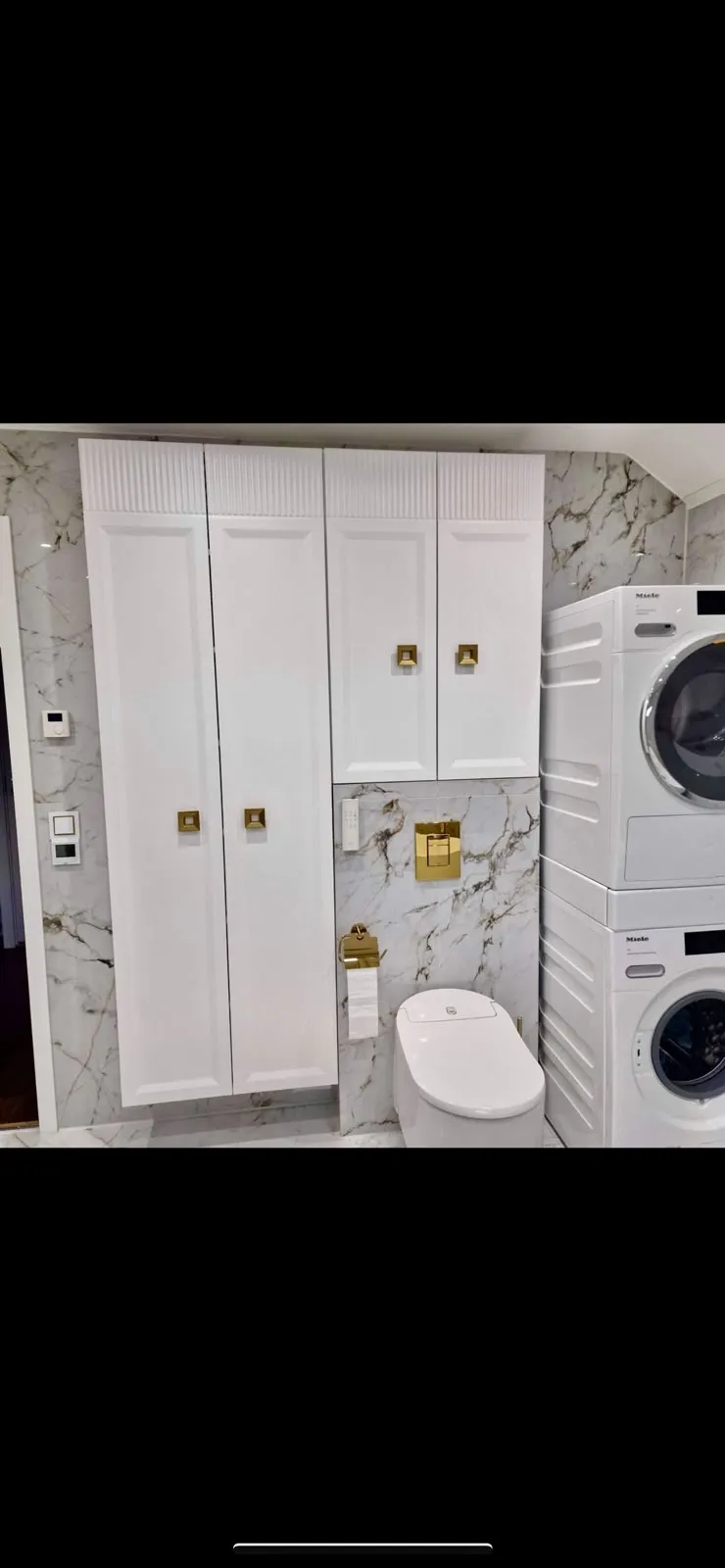 Białe meble łazienkowe nad toaletą z frezowanymi elementami dekoracyjnymi i złotymi uchwytami.